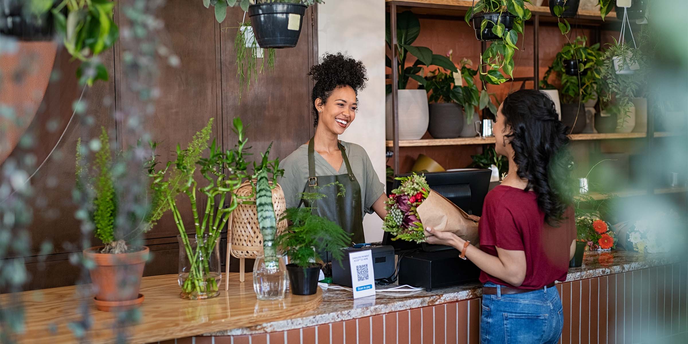 A smiling flower shop cashier warmly greets a customer purchasing a bundle of cut flowers wrapped in brown paper, representing the thriving small business success made possible through BankNewport business loans, commercial loans, and Rhode Island small business lending programs