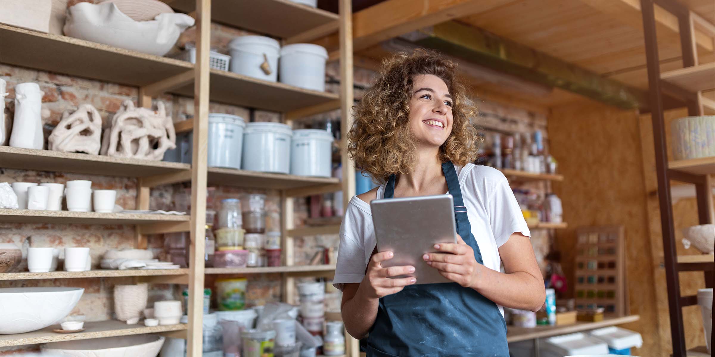 A smiling shopkeeper holds a tablet computer while standing in a storeroom filled with finished goods and materials, representing the financial preparedness and growth made possible through a business savings account, business certificate of deposit, and business CD products.