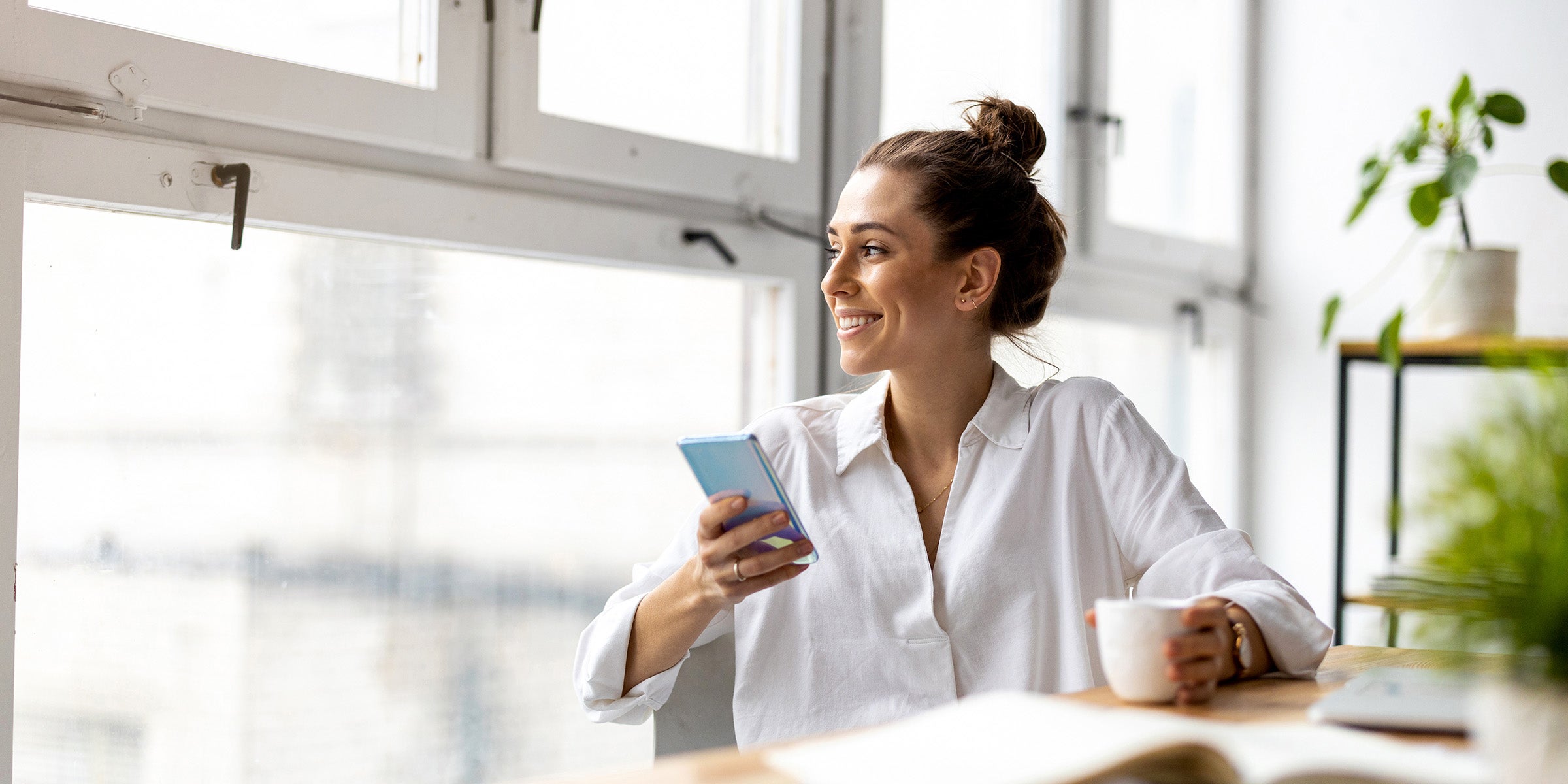 A person sits at a table in warm morning light holding a cup of coffee and a cell phone, representing the ease and accessibility of bank cash management services and payments fraud protection tools available on mobile devices.