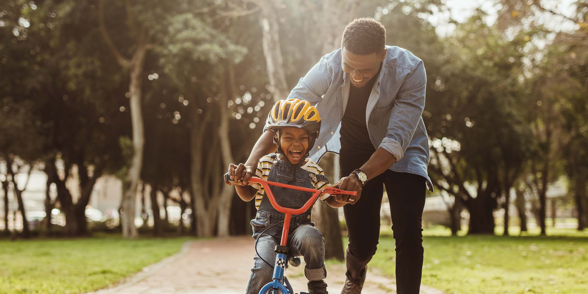 An adult helps a preschool child ride a bike along a green park path, representing the family milestones and everyday moments supported by checking and savings accounts.