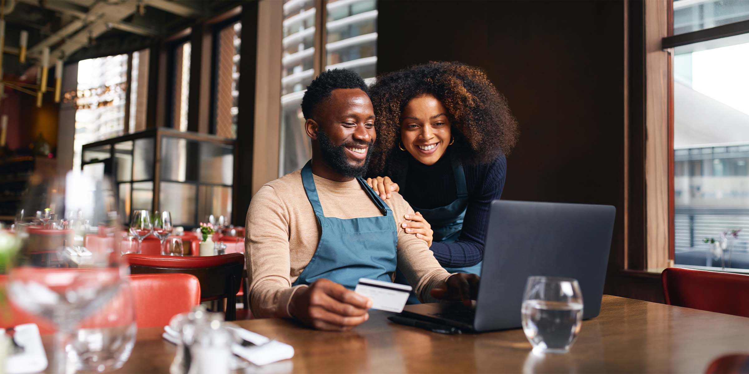 Two people sit together reviewing a laptop screen while one holds a BankNewport credit card, representing the supportive and straightforward experience of applying for a credit card — personal or business — through BankNewport's credit card program.