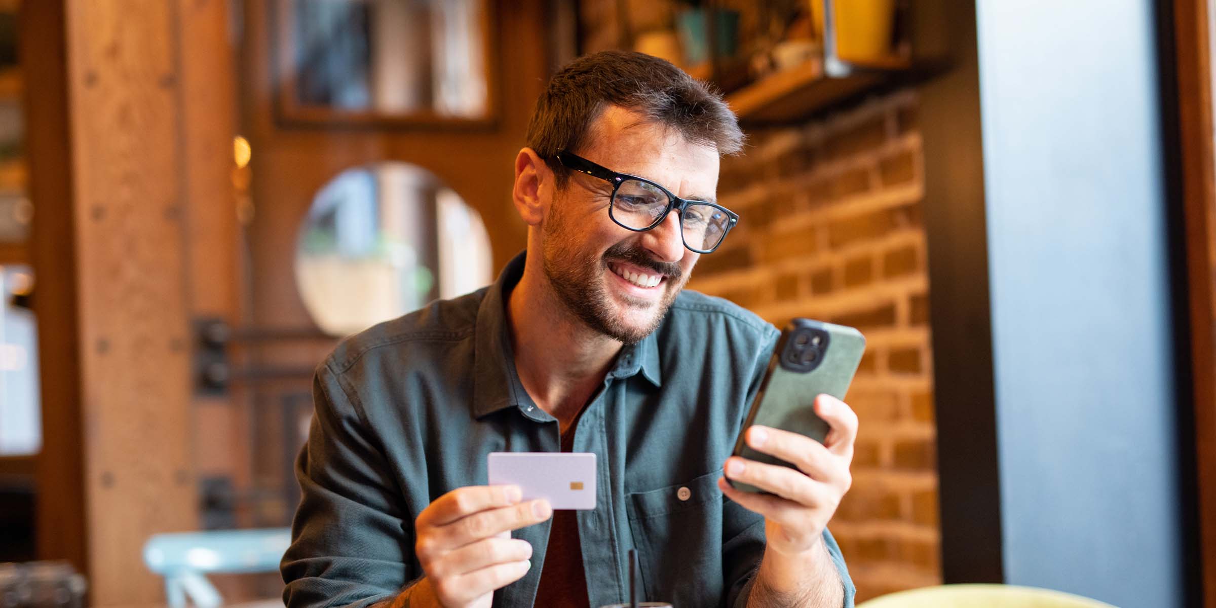 A person sits in a coffee shop holding a debit card in one hand and a mobile phone in the other, illustrating the everyday accessibility and convenience of BankNewport's digital banking services, mobile banking app, and on-the-go account management tools available to BankNewport customers.