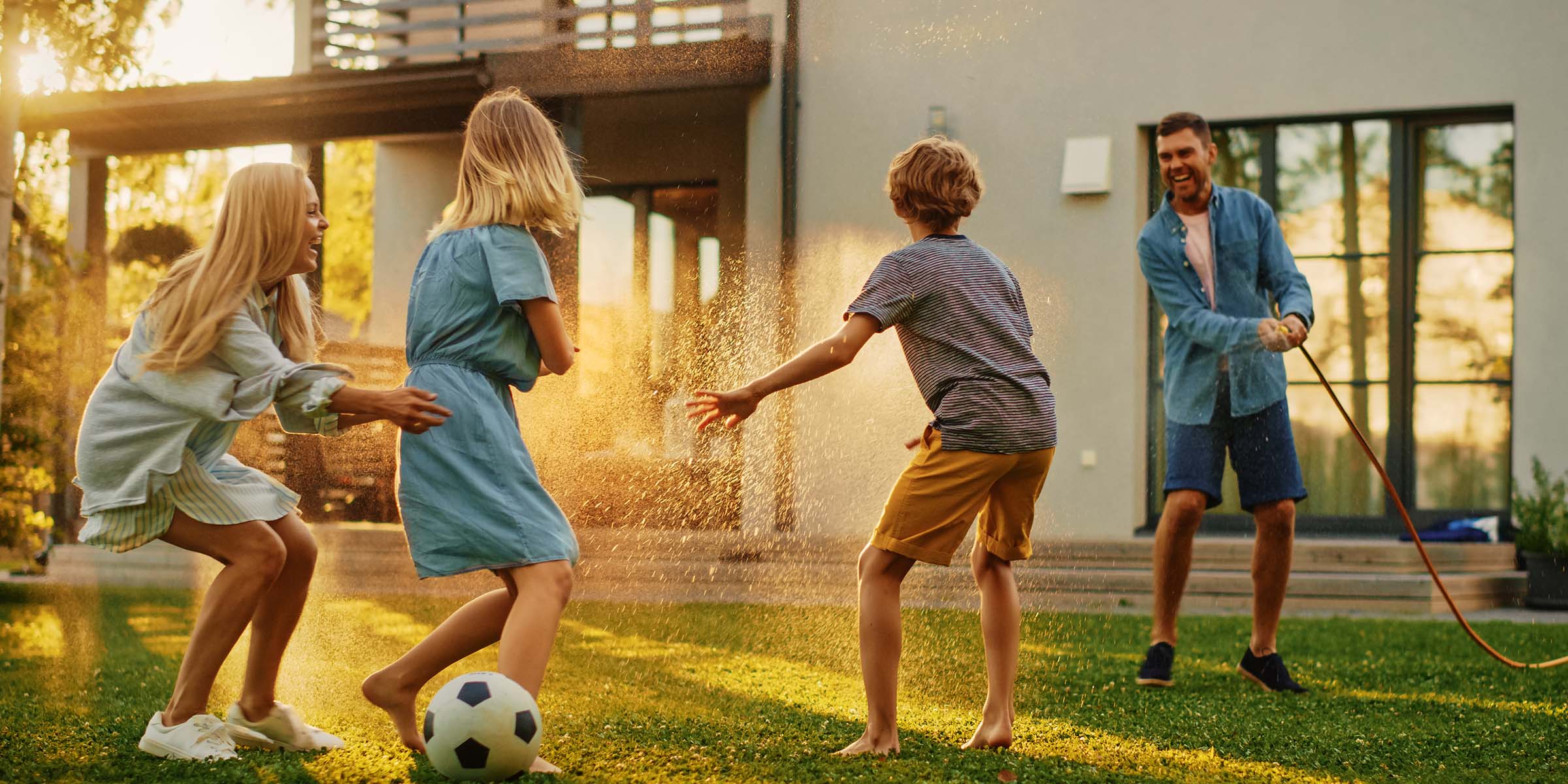 A family plays together in their backyard, laughing and dodging the spray from a garden hose as late afternoon sunlight refracts through the water droplets — representing the joyful family life and backyard moments made possible through BankNewport home loans, personal home loans, and Rhode Island home loan solutions.