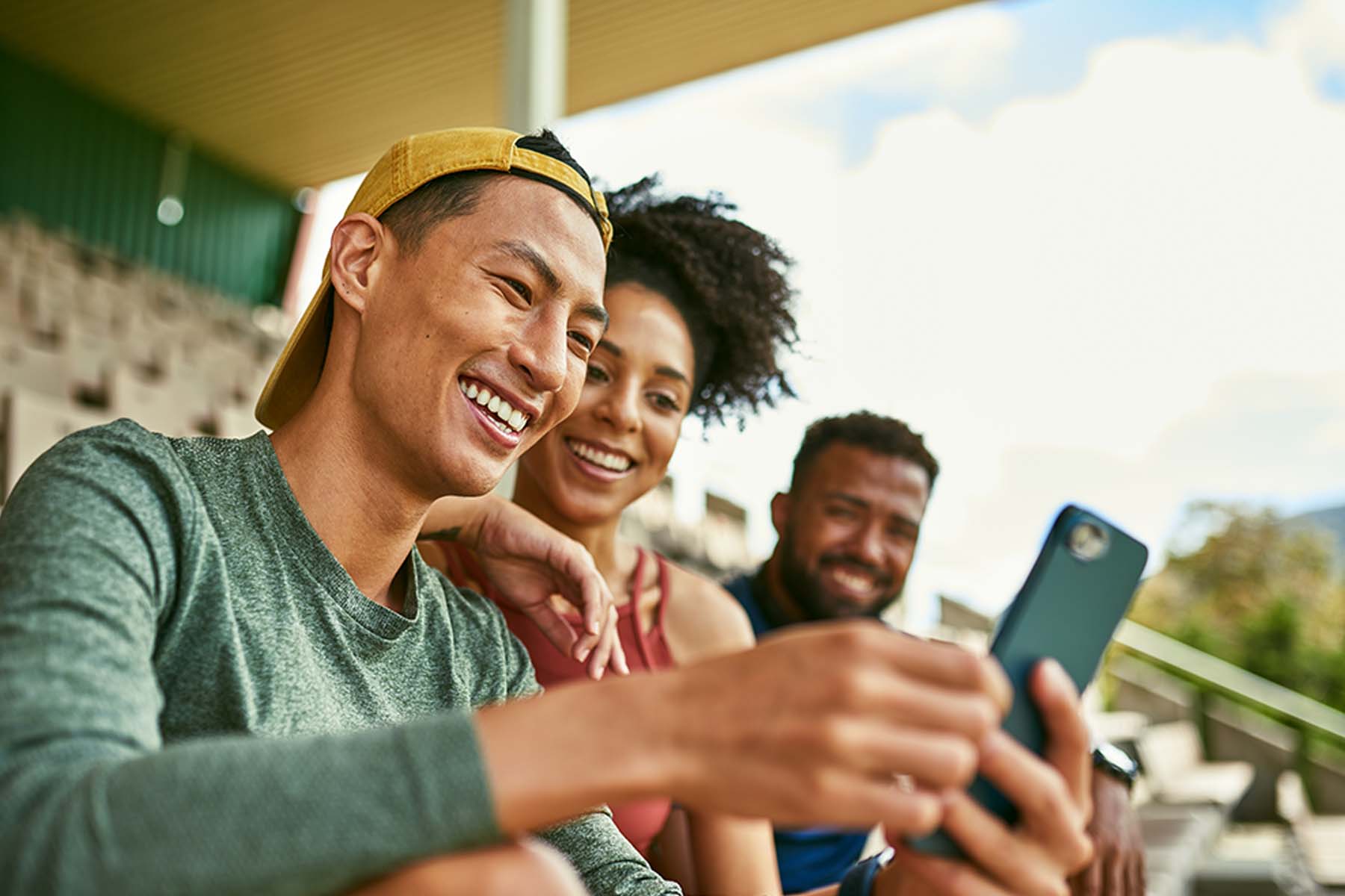 Three smiling people sit outside together looking at the screen of a cell phone held by one person, depicting the ease, accessibility, and shared convenience of digital banking and digital banking services available through BankNewport.