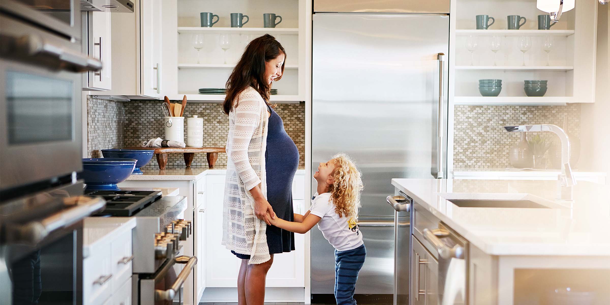 A pregnant woman gently holds hands with a young toddler in a bright, modern kitchen, representing the future-focused financial planning and family wealth building supported by BankNewport's investment services, investment solutions, guided wealth portfolios, and guided investment programs available to Rhode Island families.
