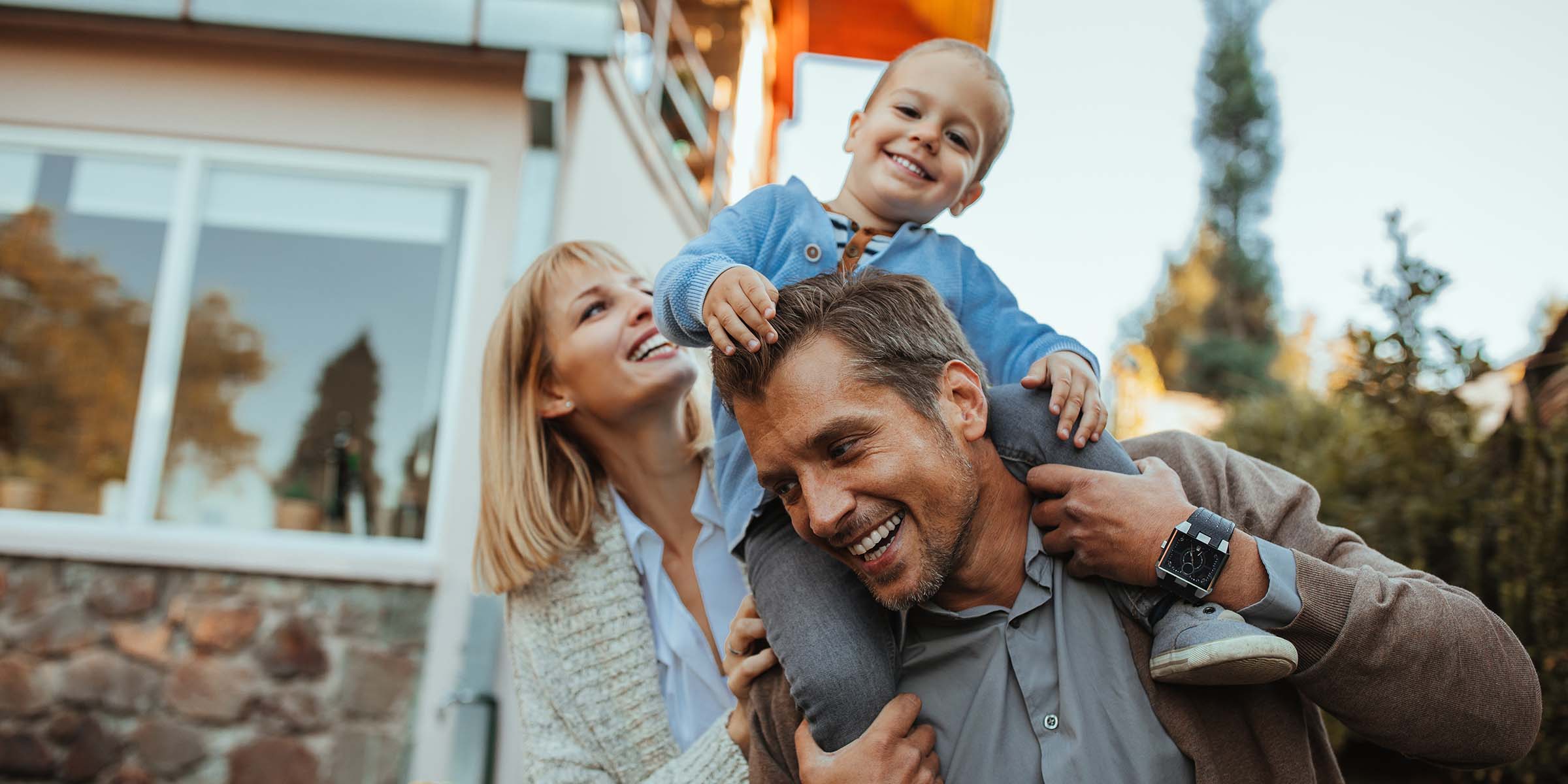 A young child rides happily on a father's shoulders as the mother looks on with relaxed, joyful smiles shared between all three — a warm family portrait representing the milestone of homeownership made possible through BankNewport mortgage lending in Rhode Island