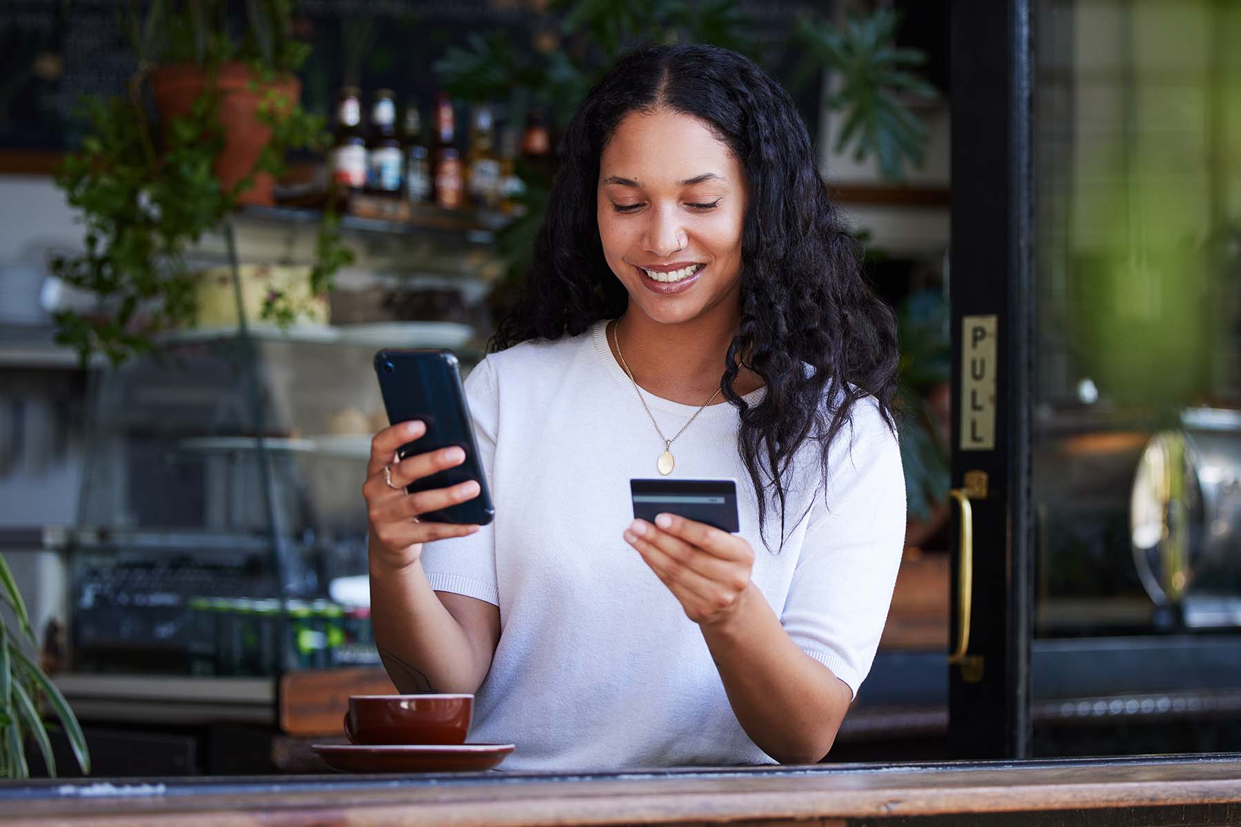 A person sits in a coffee shop examining a debit card in one hand and a mobile phone in the other, representing the everyday financial awareness and accessibility of opening and managing a personal savings account with BankNewport